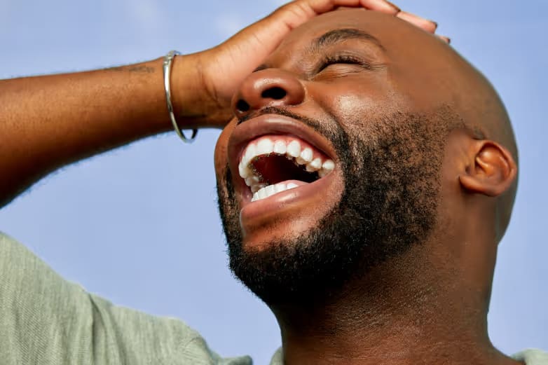 Man with straight, white teeth, looking into the sky, one hand on his head, smiling broadly.