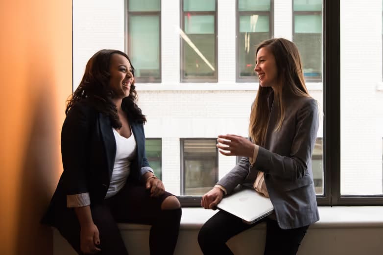 Two women sitting on the window sill of a professional building talking.